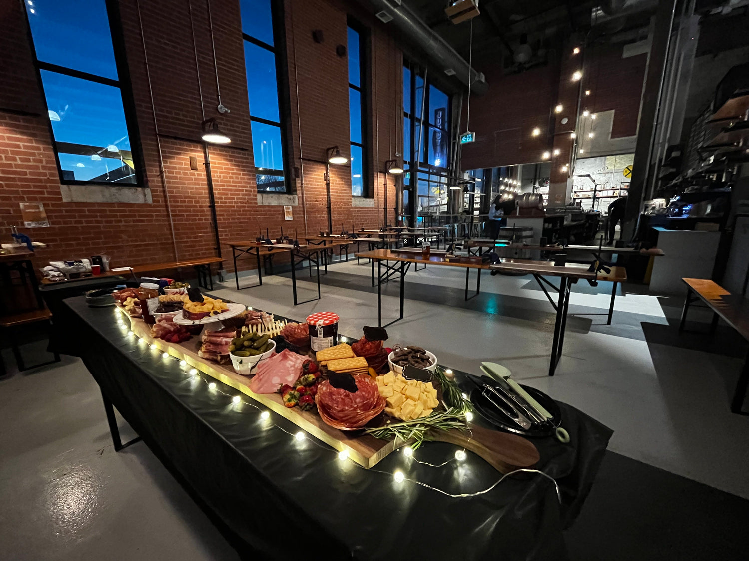 a beautifully decorated large charcuterie board loaded with meats, cheeses, fruits and more. It is surrounded by lights.  There is a setup of 20 stations waiting for people to arrive and begin the charcuterie board workshop.