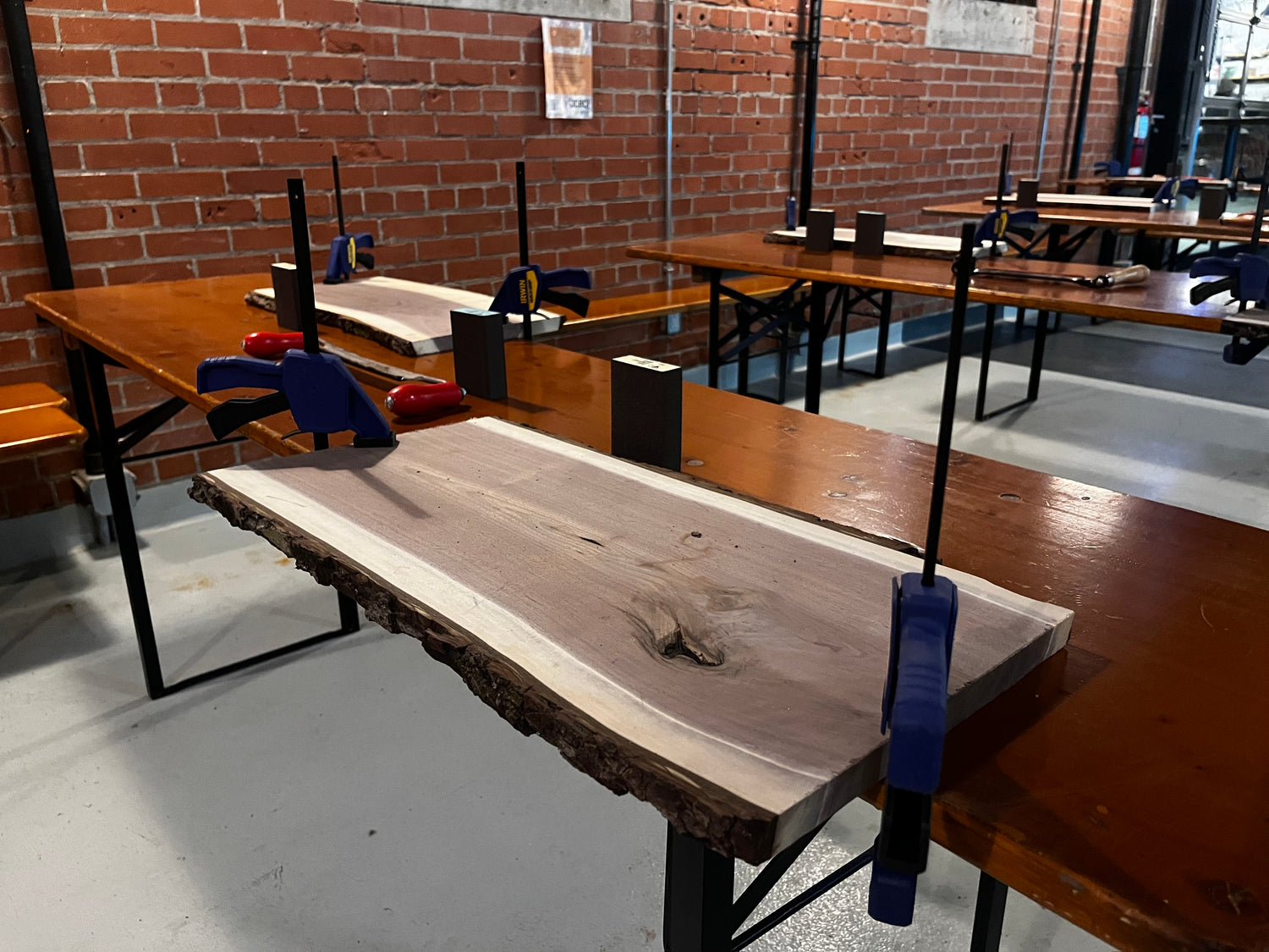 The setup before an event begins. Each station has a 24" slab of walnut wood held down by two clamps, a draw knife and a sanding block.  There are about 15 stations set up in a brewery with a brick wall and windows.